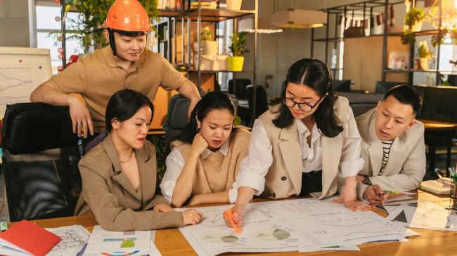 Diverse team of five professionals collaboratively working on architectural plans and financial charts spread out on a large table.