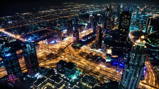 Aerial night view of a bustling metropolis with illuminated skyscrapers and busy traffic interchanges.