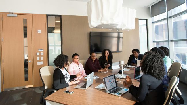 A diverse group of professionals sitting around a conference table discussing sustainable urban drainage systems in a well-lit, modern office setting with laptops and digital devices visible.