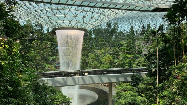 A lush indoor garden featuring a large central water fountain under a geometric glass dome, showcasing water reuse and management strategies.